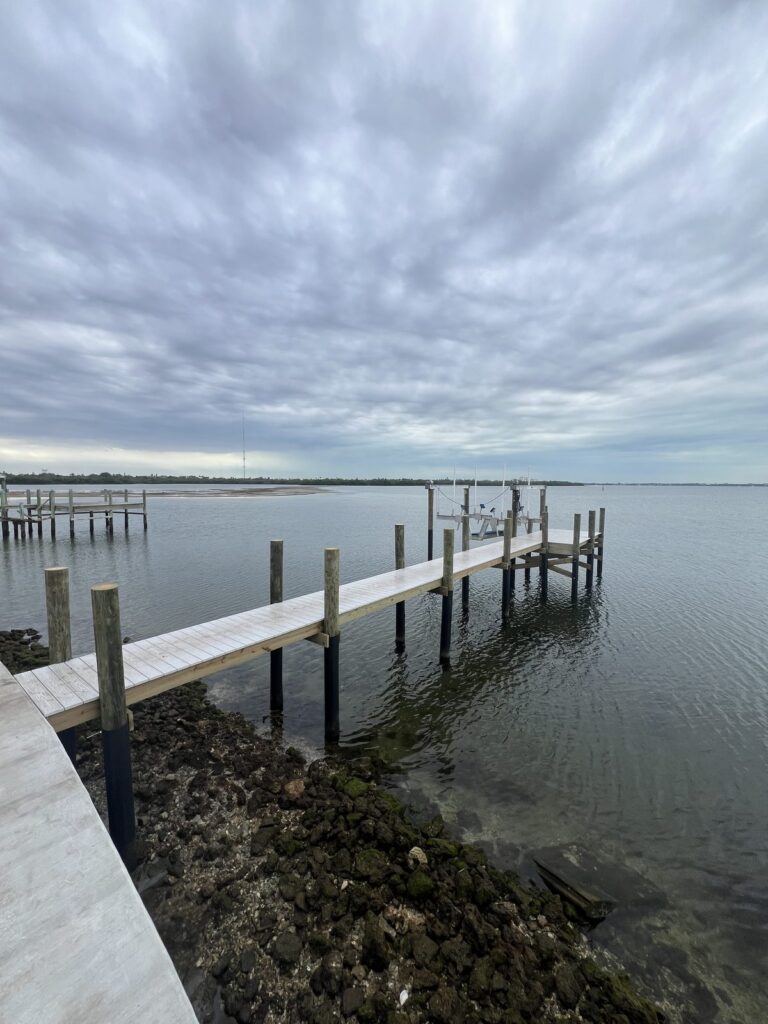 Finished concrete seawall topped with a wood cap, riprap rock along the base, and wooden pilings with black wraps at the waterline standing in front of the wall. Seawall installation by Anderson Dock and Lift, Bradenton FL.